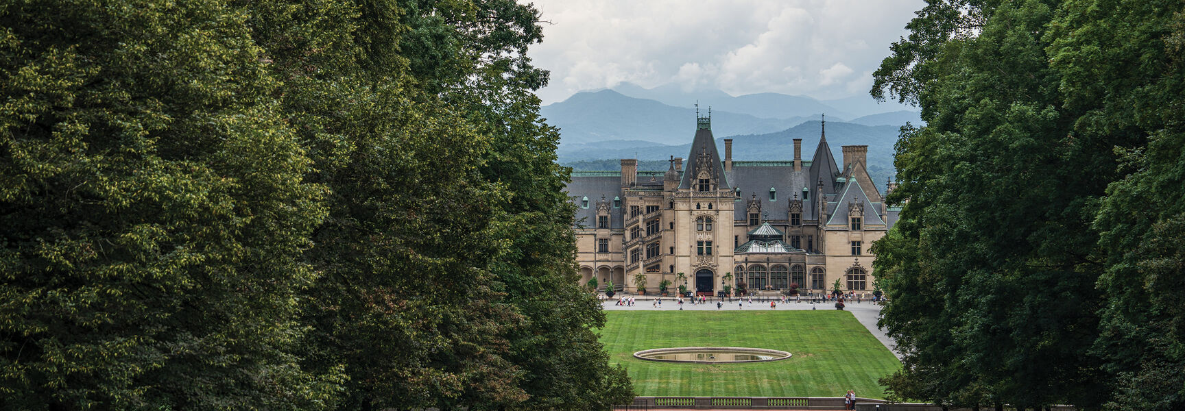 The historic Biltmore Estate in Asheville, North Carolina, framed by lush green trees with a vast lawn and mountain peaks in the distance.