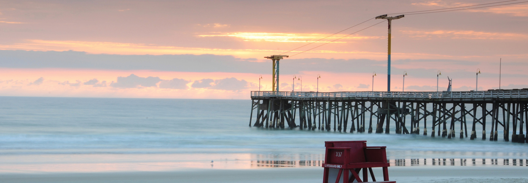 A red lifeguard chair sits on the beach in front of a long pier during a tranquil sunset in Key West, Florida.
