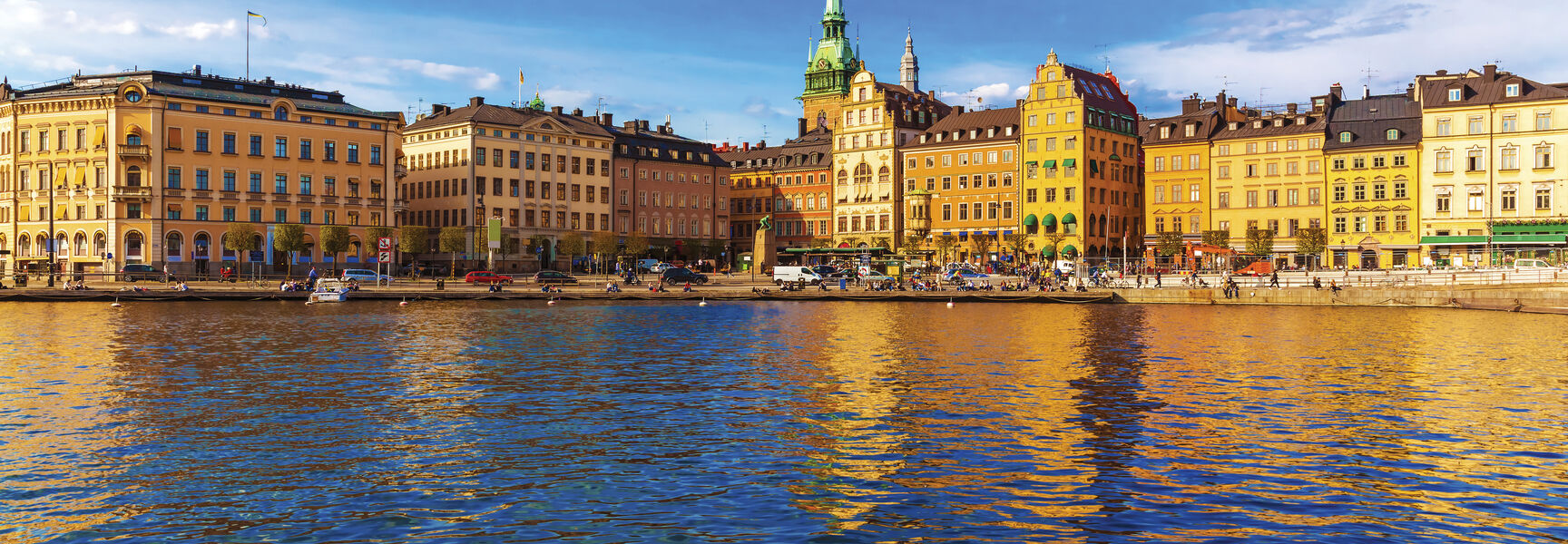 Vibrant historic buildings and a green church spire along the Stockholm waterfront reflect in the blue harbor water under a bright, cloudy sky.