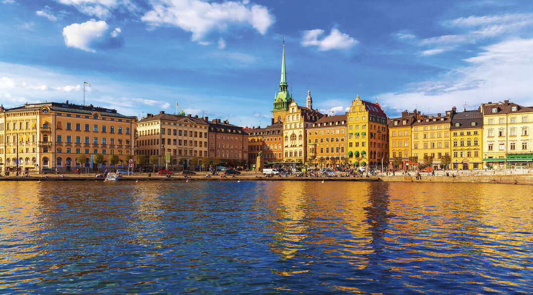 Vibrant historic buildings and a green church spire along the Stockholm waterfront reflect in the blue harbor water under a bright, cloudy sky.