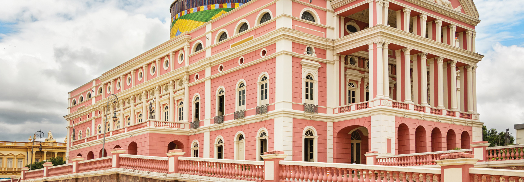 The pink and white Amazon Theatre, an ornate opera house, stands under a cloudy sky in Manaus, Brazil, with its colorful dome partially visible.