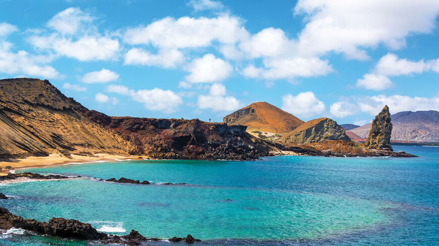 Volcanic landscape in the Galápagos Islands featuring turquoise water, rocky cliffs, and a prominent rock pinnacle under a blue sky with white clouds.