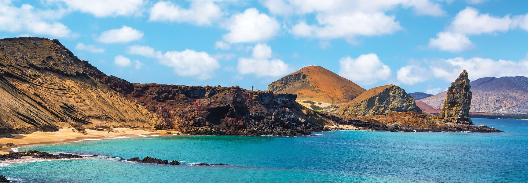 Volcanic landscape in the Galápagos Islands featuring turquoise water, rocky cliffs, and a prominent rock pinnacle under a blue sky with white clouds.