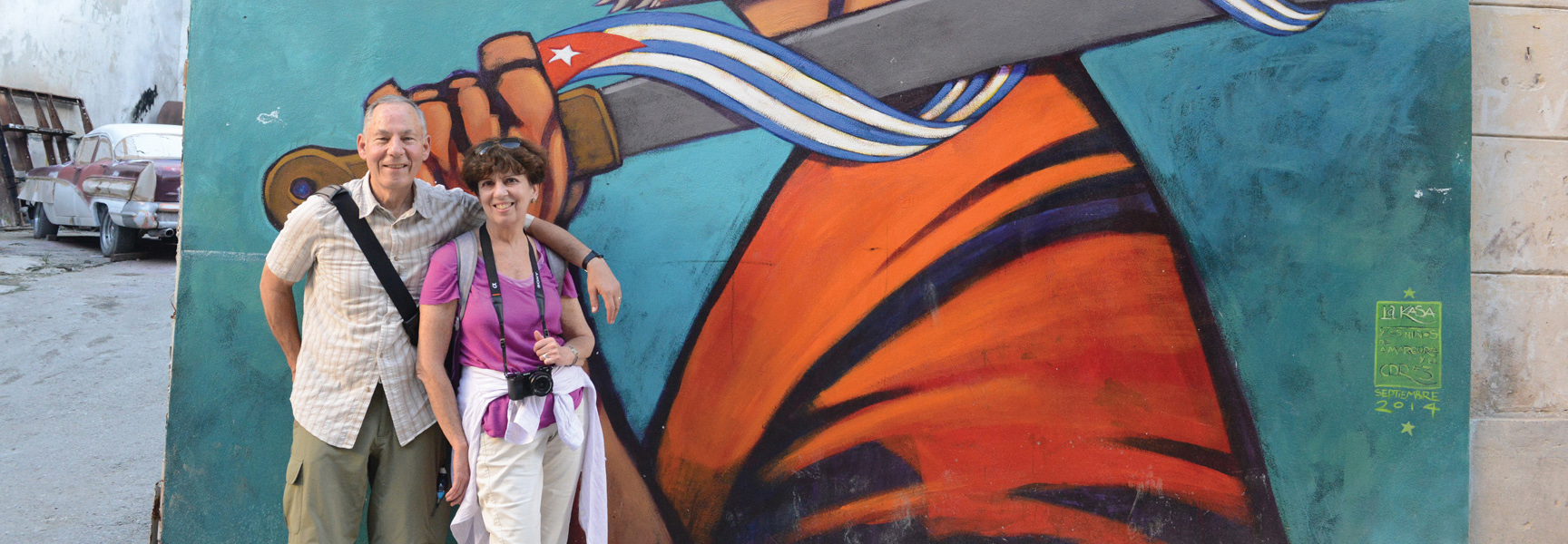 A couple poses for a photo in front of a colorful mural and a classic car on a street in Cuba.