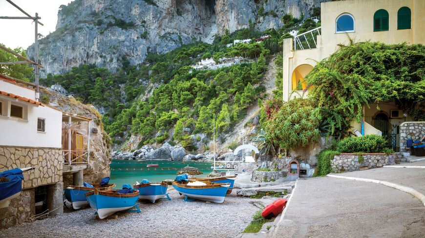 Small blue boats rest on a pebbled beach in Sorrento, Italy, with turquoise water and lush green cliffs rising behind coastal buildings.