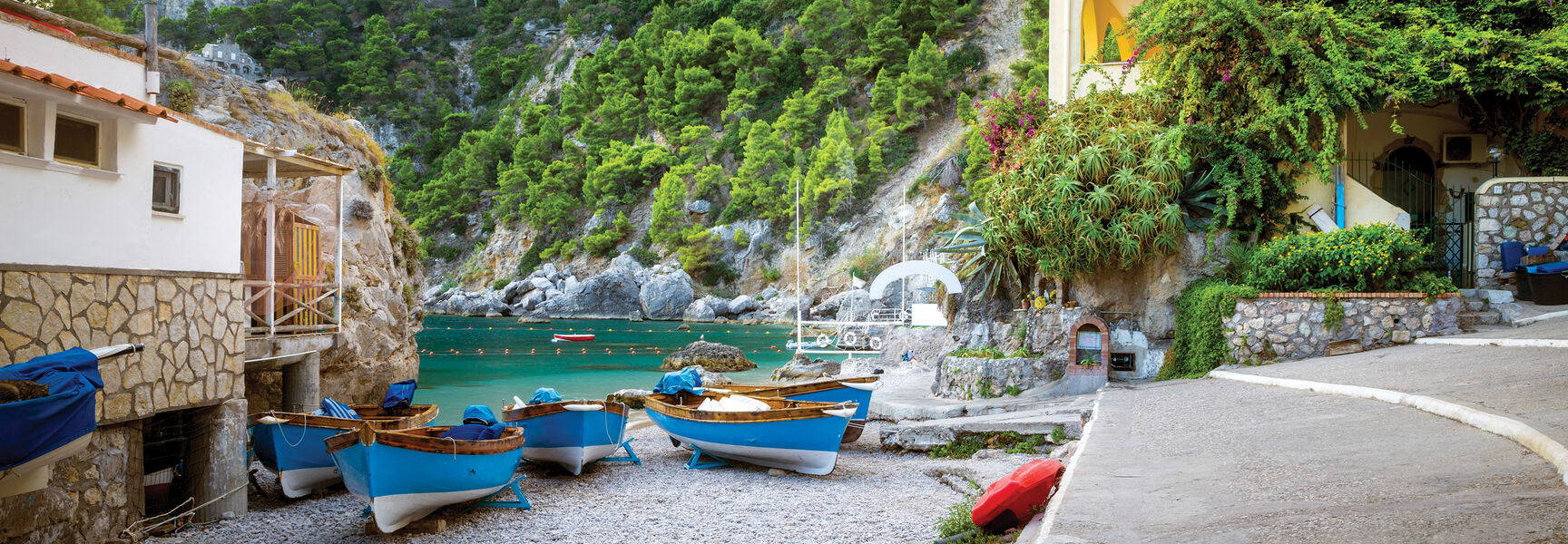 Small blue boats rest on a pebbled beach in Sorrento, Italy, with turquoise water and lush green cliffs rising behind coastal buildings.