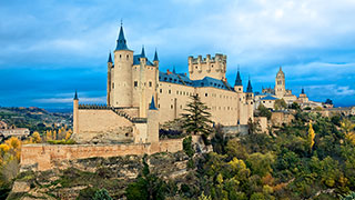 The Alcázar Castle in Spain sits on a tree-covered hill against a partly cloudy blue sky.