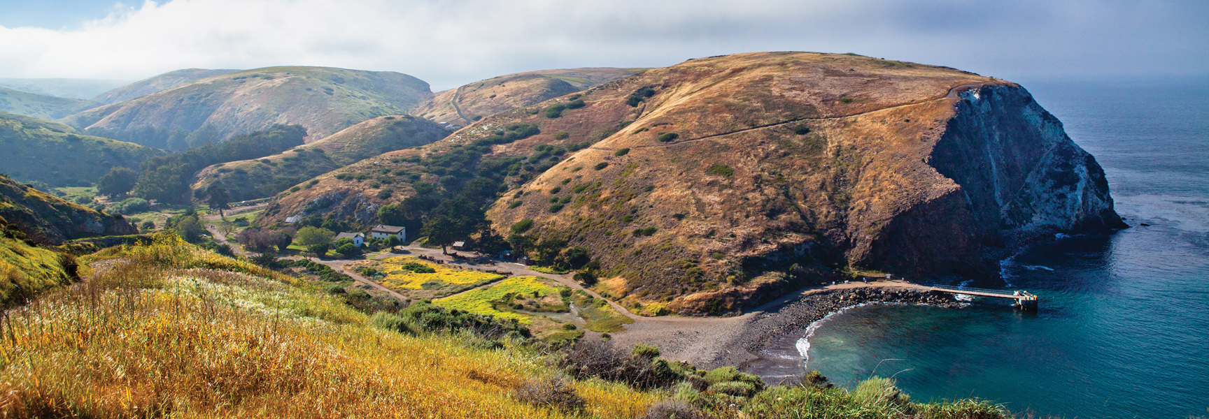 A wide view of the hilly, golden-brown Channel Islands in California, with a pier extending into a small blue cove.