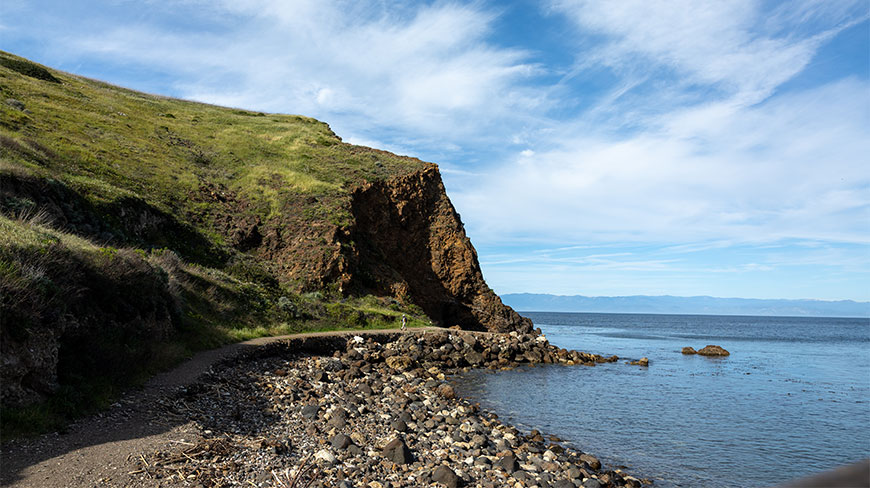 A person walks along a coastal path below a steep grassy cliff on Santa Cruz Island in California's Channel Islands National Park.