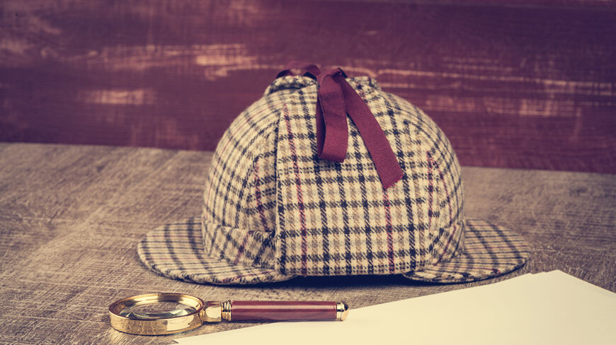 A Sherlock Holmes style deerstalker hat and magnifying glass rest on a wooden table beside blank paper, representing Sherlock Holmes’ legacy and mystery.