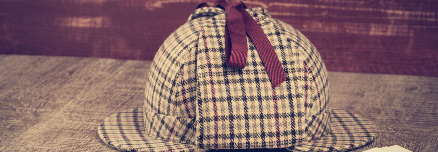 A Sherlock Holmes style deerstalker hat and magnifying glass rest on a wooden table beside blank paper, representing Sherlock Holmes’ legacy and mystery.