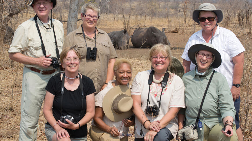 A group of smiling travelers on a Botswana safari with two rhinoceroses resting in the dry wilderness landscape behind them.