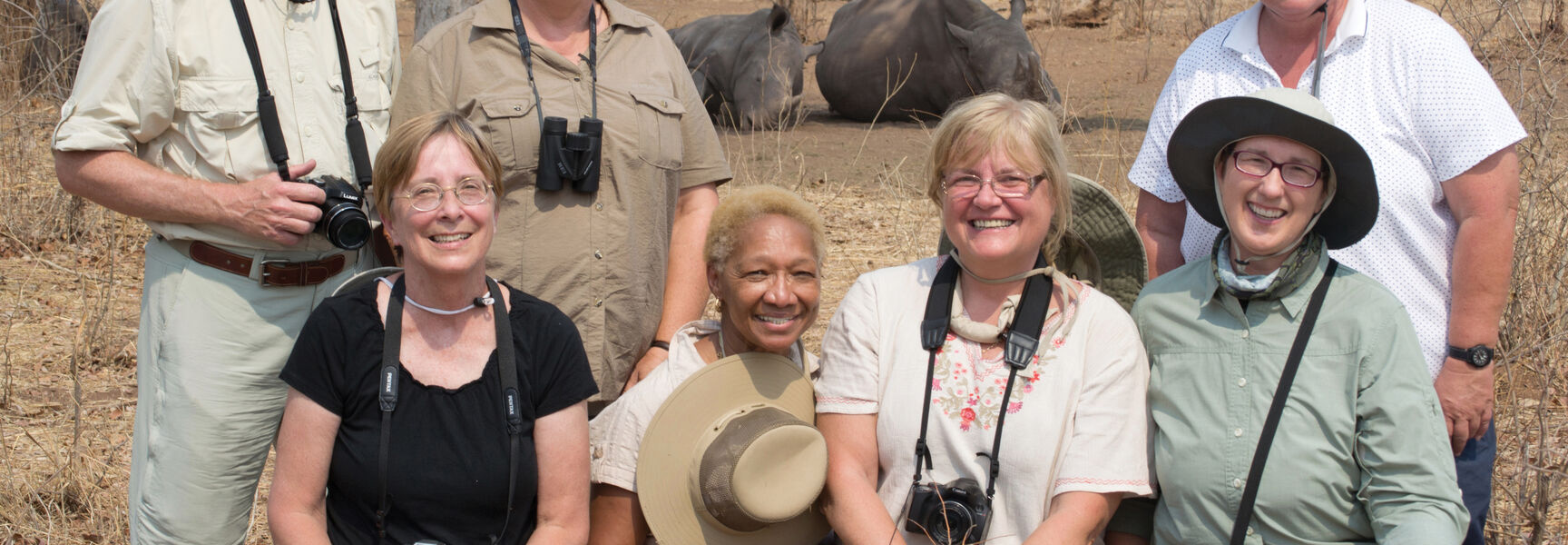 A group of smiling travelers on a Botswana safari with two rhinoceroses resting in the dry wilderness landscape behind them.