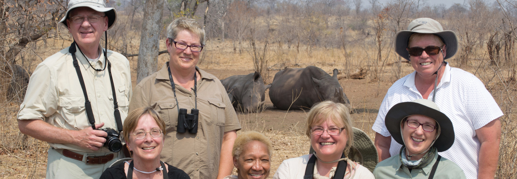A group of seven travelers on safari in Botswana smile for a photo with two large rhinos resting in the background.