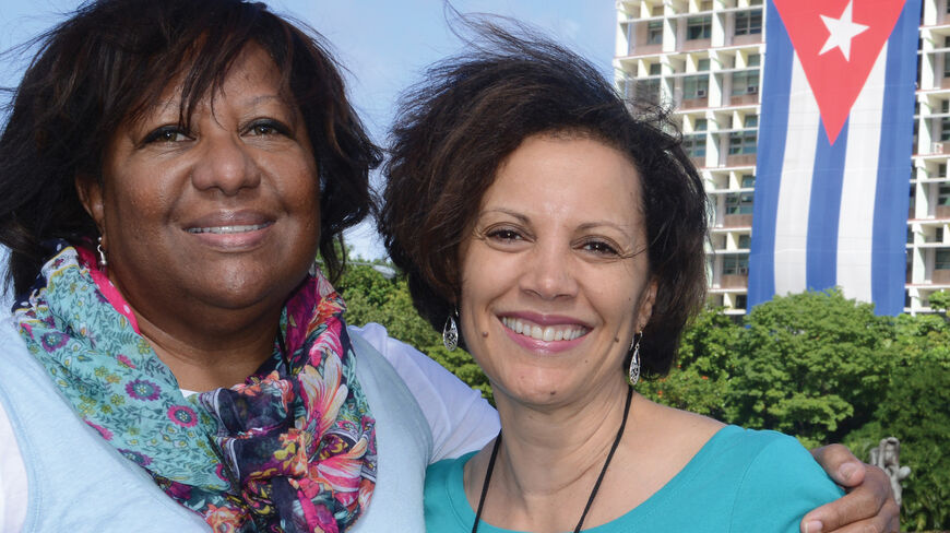 Two women smile in front of a building draped with a large Cuban flag during a Road Scholar walking adventure in historic Havana, Cuba.