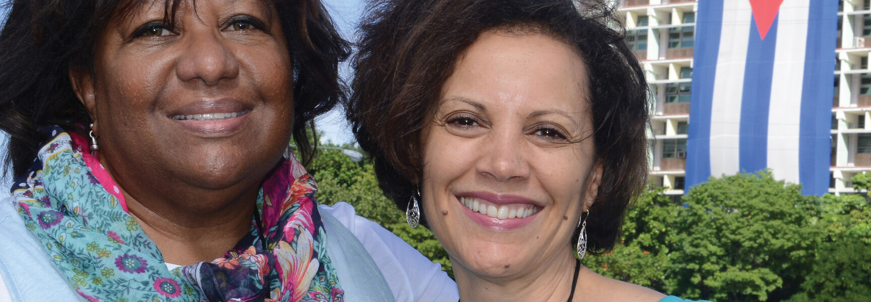 Two women smile in front of a building draped with a large Cuban flag during a Road Scholar walking adventure in historic Havana, Cuba.