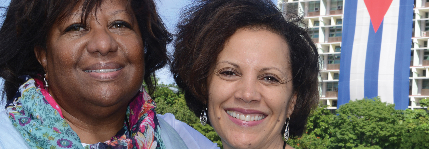 Two smiling women in Cuba with the Cuban flag in the background.