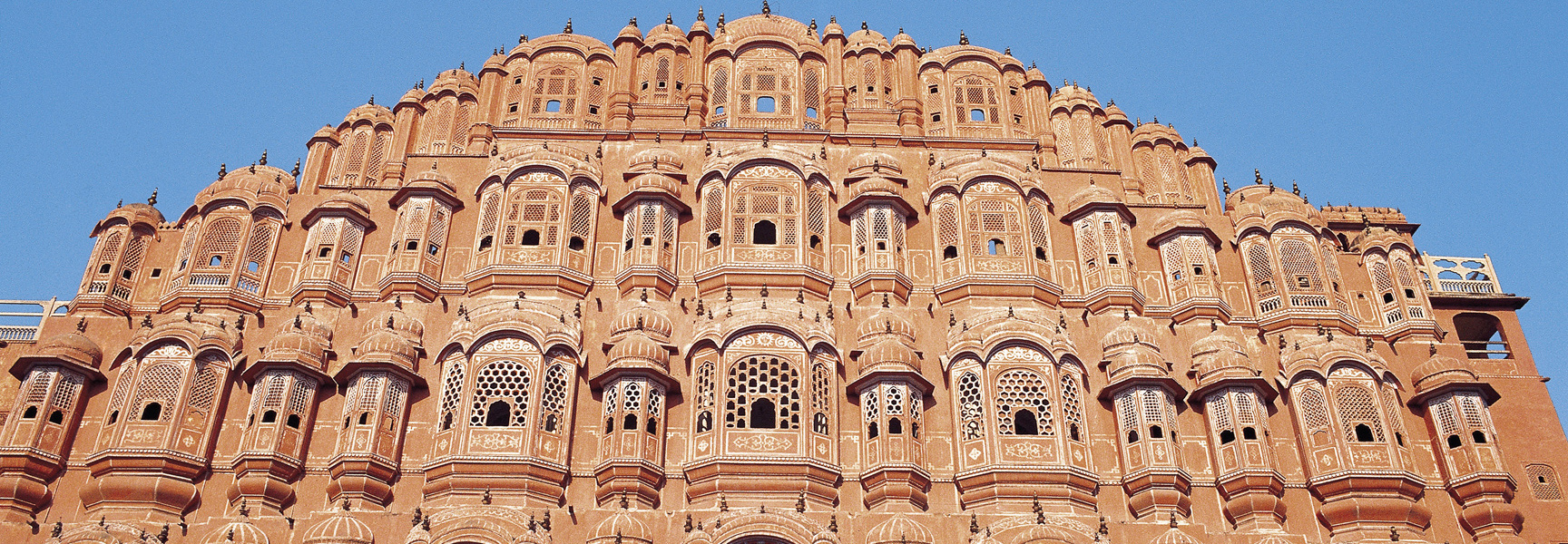 The intricate pink sandstone facade of the Hawa Mahal palace in Jaipur, India, shown against a bright blue sky.