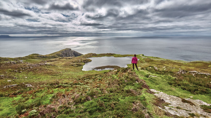 A hiker in a pink jacket stands on a grassy hill overlooking a lake and the sea along Ireland’s coastal northwest under cloudy skies.