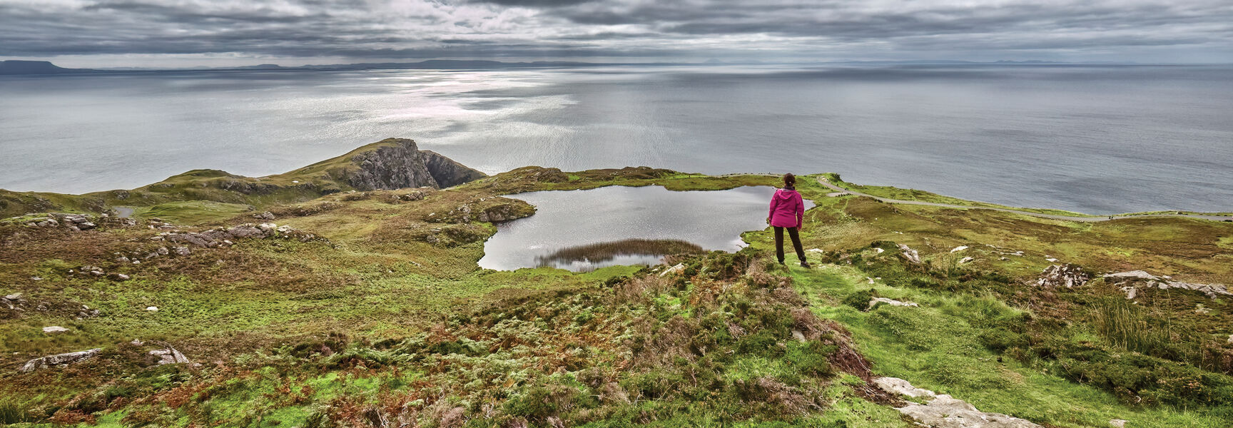 A hiker in a pink jacket stands on a grassy hill overlooking a lake and the sea along Ireland’s coastal northwest under cloudy skies.