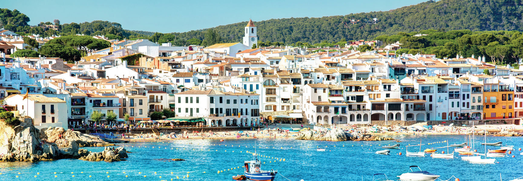 Whitewashed buildings of a coastal village in Spain overlook a bright blue bay with boats and a sandy beach full of people.