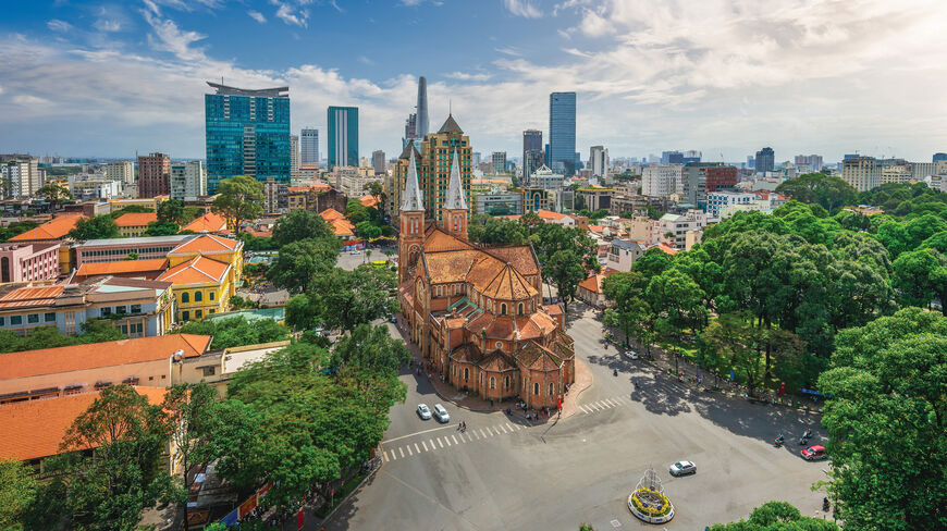 Aerial view of the Notre-Dame Cathedral Basilica of Saigon surrounded by lush green trees and modern skyscrapers in Ho Chi Minh City, Vietnam.