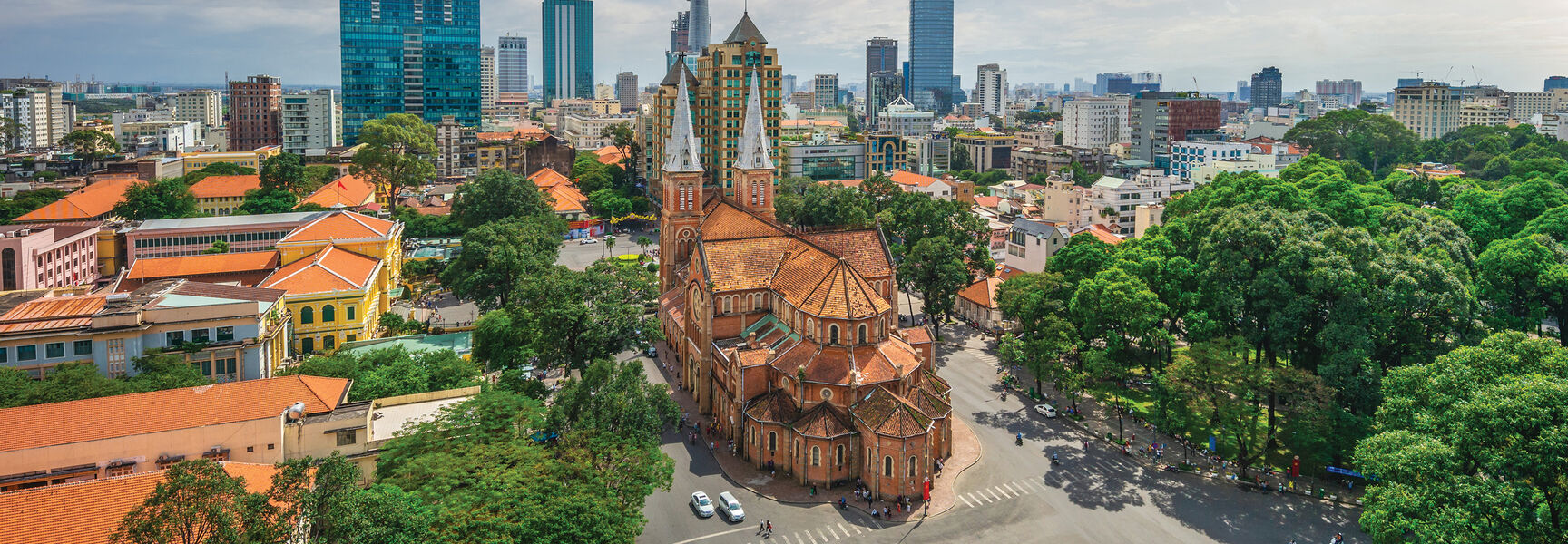 Aerial view of the Notre-Dame Cathedral Basilica of Saigon surrounded by lush green trees and modern skyscrapers in Ho Chi Minh City, Vietnam.