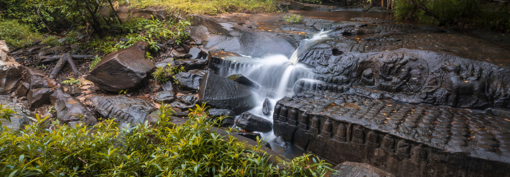 A small waterfall cascades over ancient Khmer stone carvings in a riverbed surrounded by lush green plants in Kbal Spean, Cambodia.
