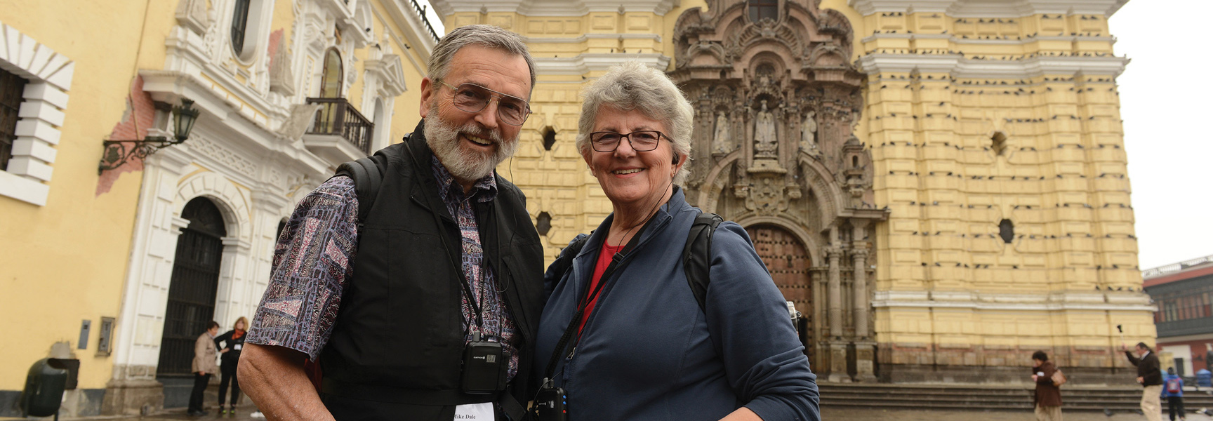 A smiling couple stands in front of a large, historic, yellow building in Peru.