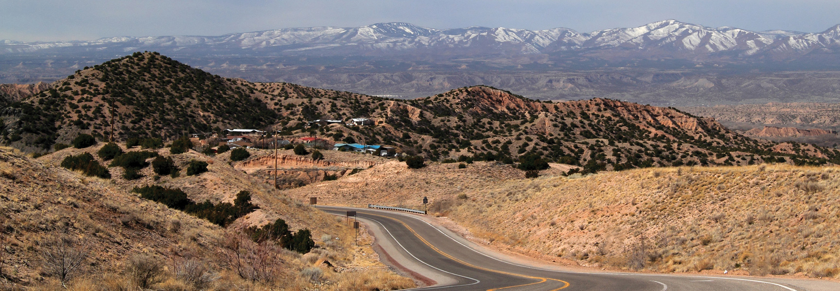 A winding road runs through the hilly landscape of New Mexico, with snow-capped mountains visible in the distance.