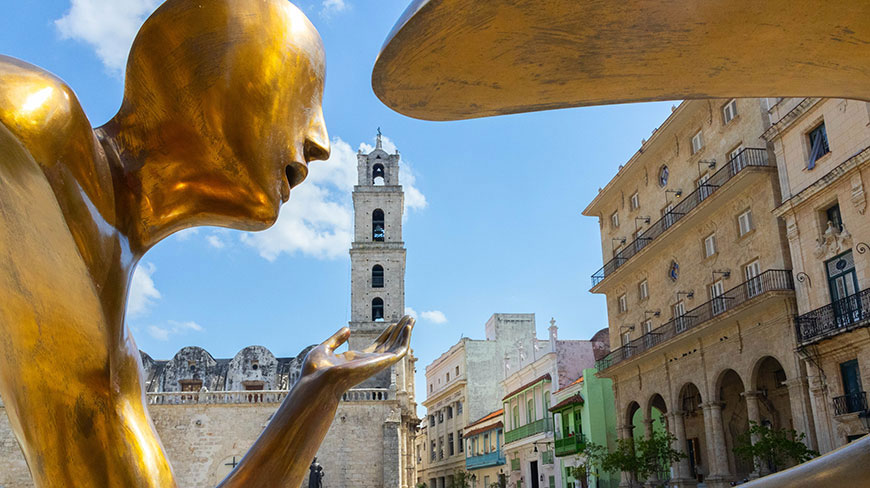A golden statue in Plaza de San Francisco in Havana, Cuba, frames a historic bell tower in the background under a blue sky.