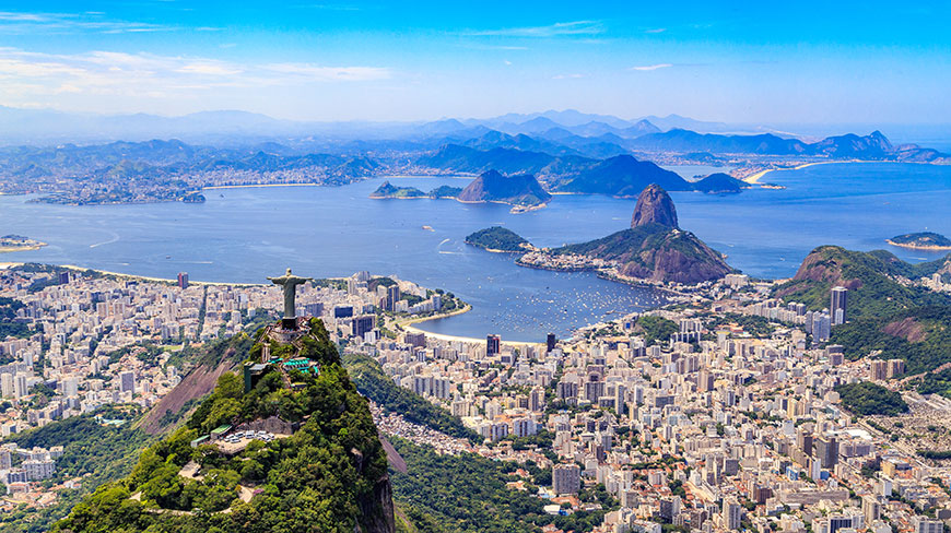 An aerial view of Rio de Janeiro, Brazil, shows the Christ the Redeemer statue overlooking the city, Sugarloaf Mountain, and the bay.
