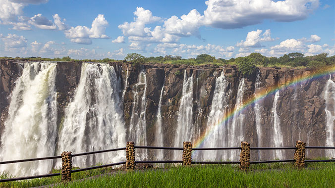 A wide, scenic view of Victoria Falls in Africa, with a bright rainbow arching through the mist in front of the cascading water.