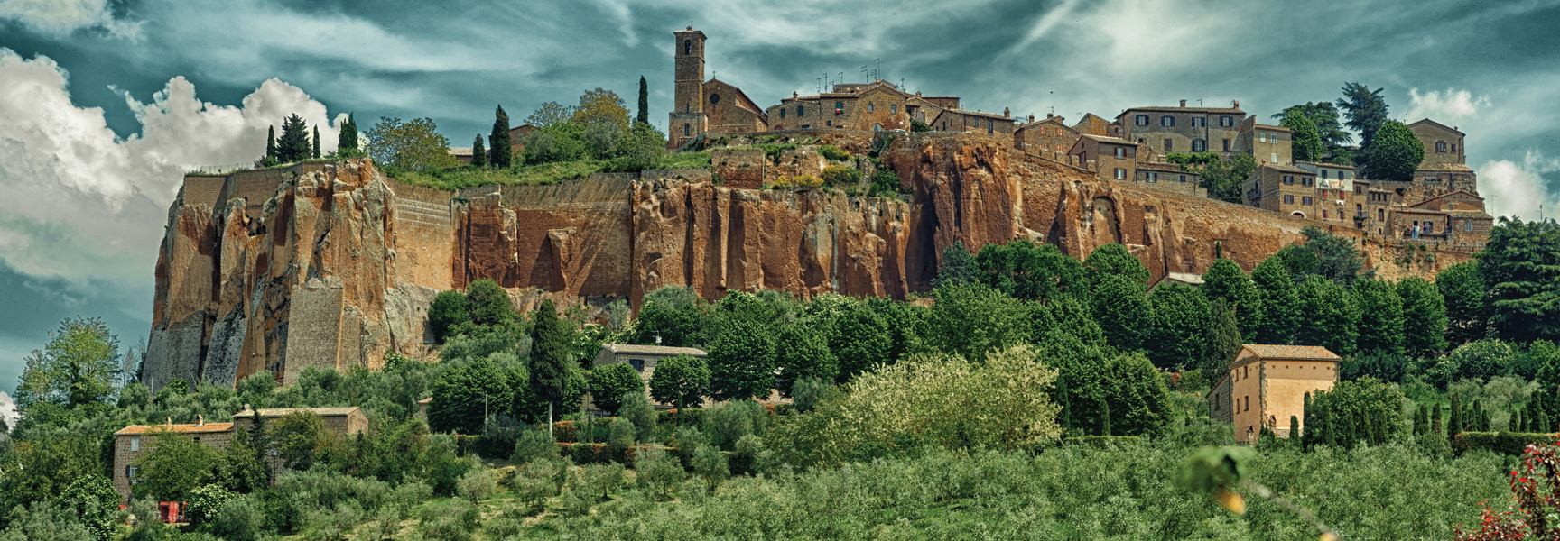 A historic Italian village sits atop a dramatic, steep cliff, overlooking a lush valley of green trees under a partly cloudy sky.