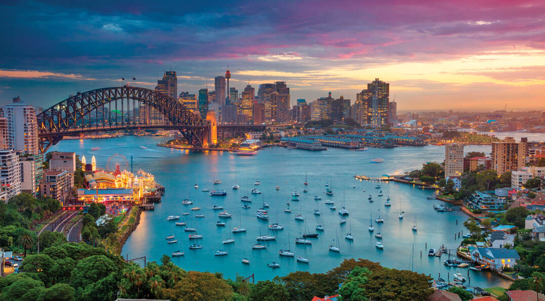 A vibrant sunset over the Sydney Harbor Bridge and the illuminated city skyline, with numerous boats anchored in the harbor below a colorful sky.