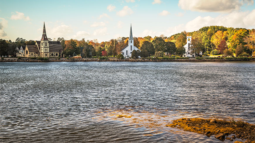 The Three Churches of Mahone Bay stand along the waterfront in Nova Scotia, surrounded by colorful autumn foliage under a blue sky.