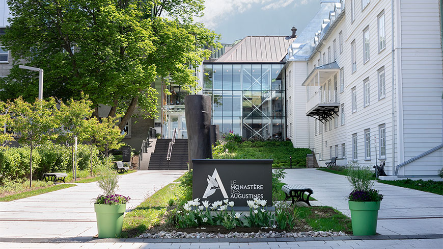 The paved walkway and entrance to Le Monastère des Augustines in Québec, showing a mix of modern and historic architecture surrounded by leafy green trees.