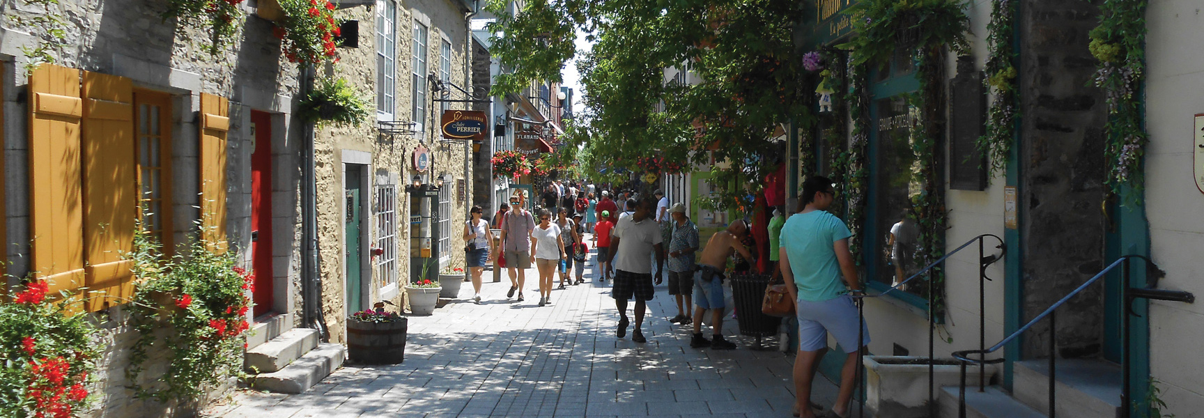 A crowd of people walks down a sunny, historic cobblestone street in Québec, lined with old stone buildings and shops.