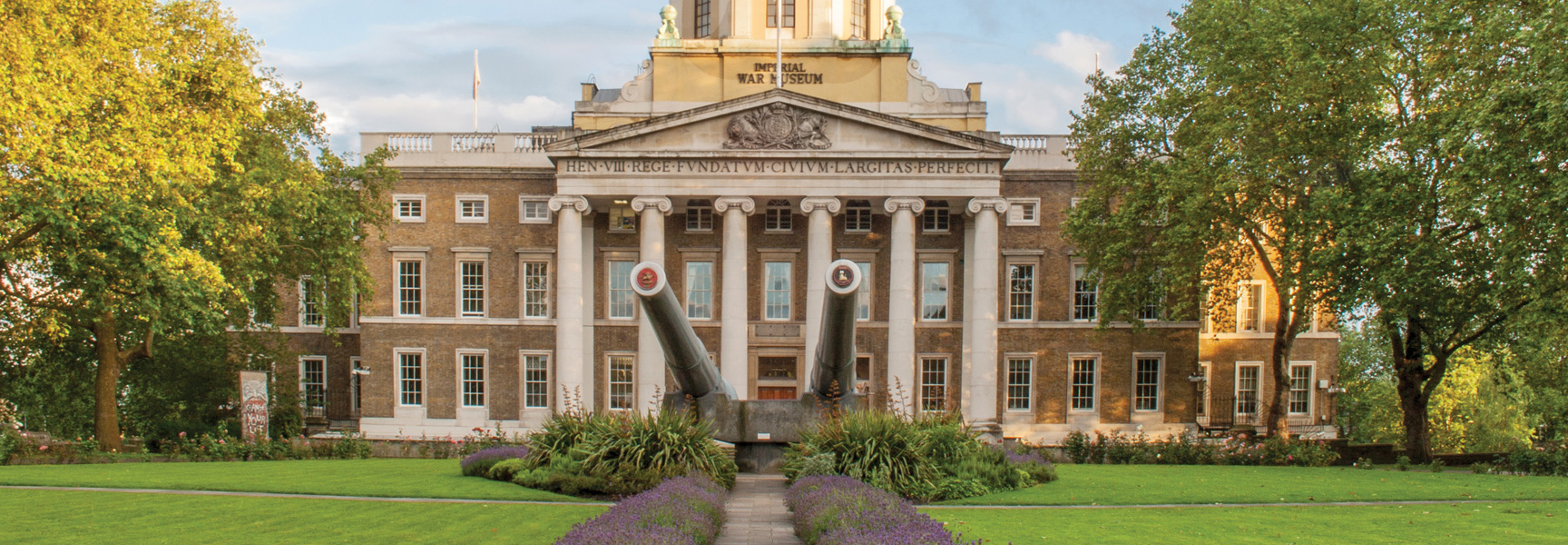 The grand entrance to the Imperial War Museum in England, with two large naval guns displayed on the front lawn under a golden sky.