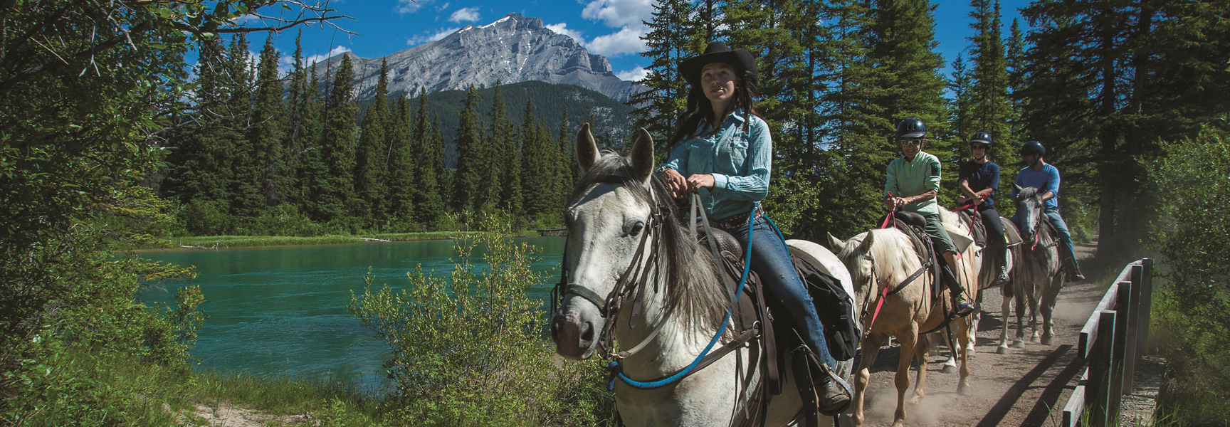 A family horseback rides on a trail next to a turquoise river in the Canadian Rockies in Alberta.