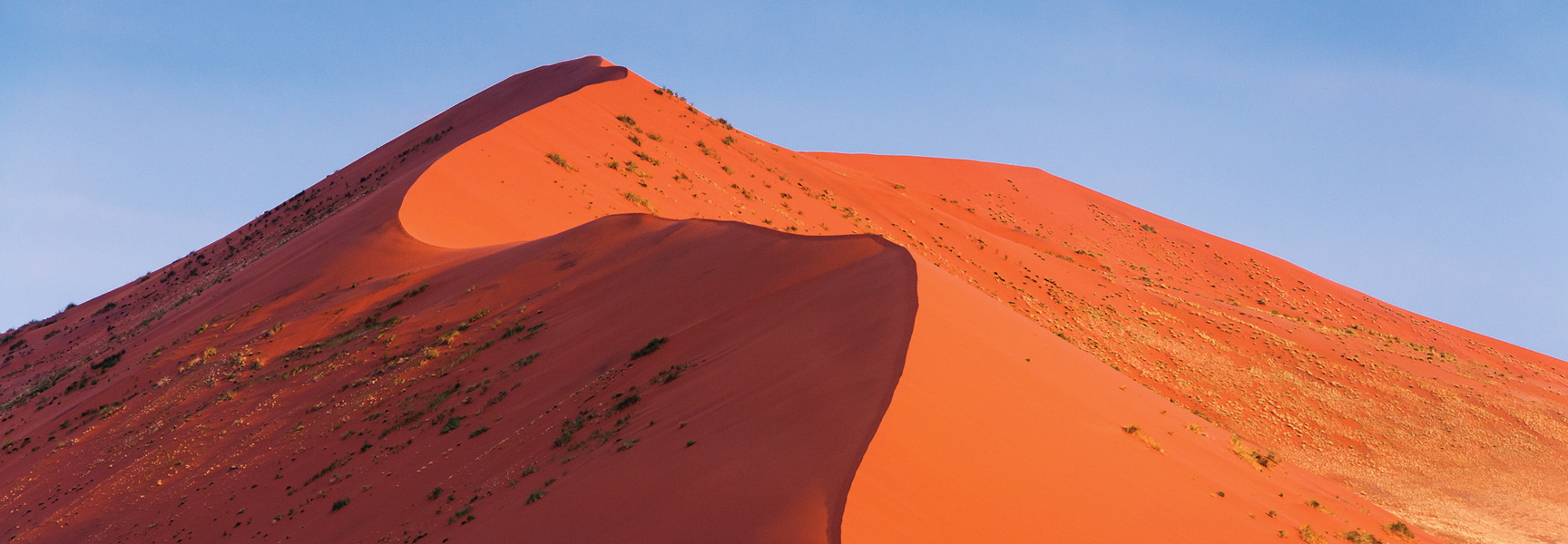The sun illuminates a massive red sand dune with a curved crest against a clear blue sky in Africa.