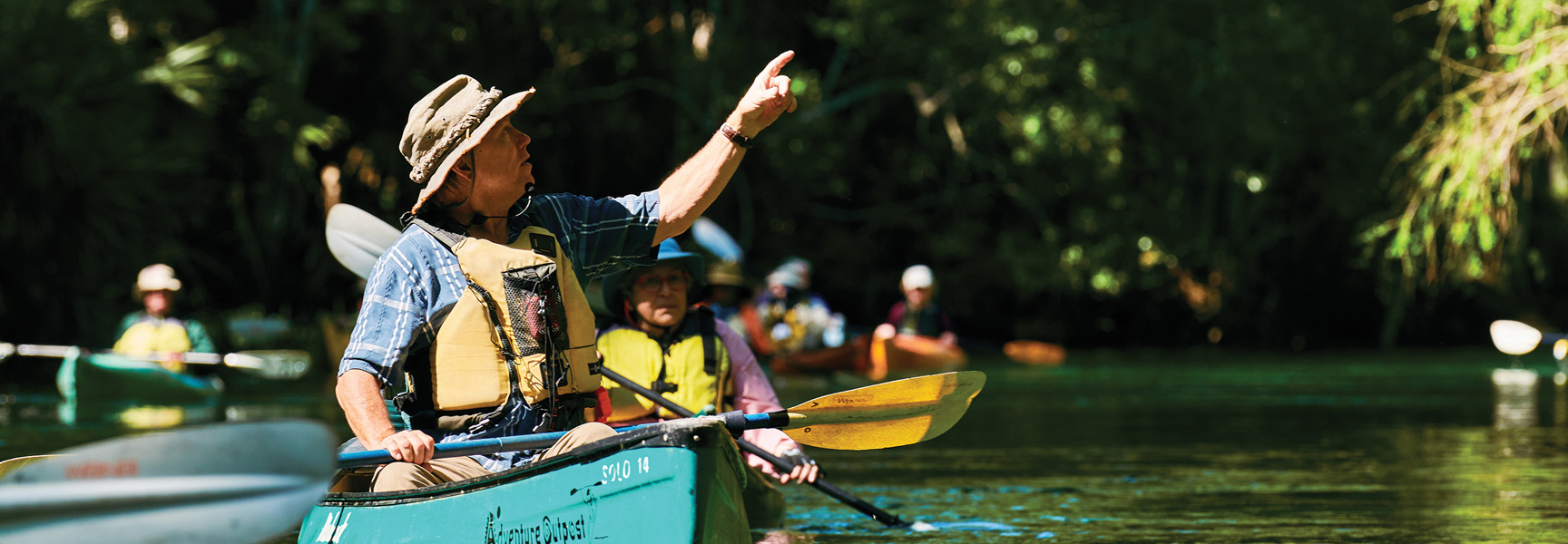 A man in a hat and life vest points forward while leading a group of people kayaking on a river in Florida.