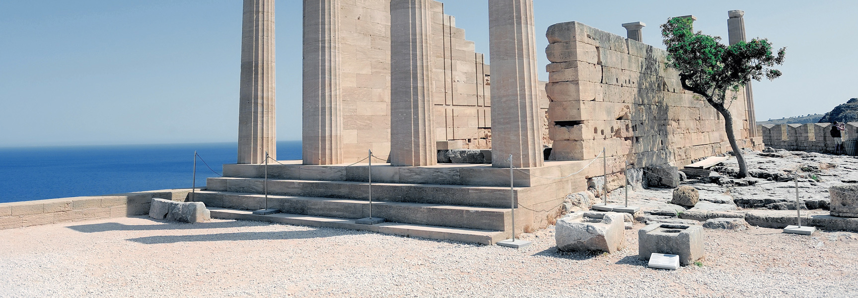 Ancient Greek ruins with large stone columns stand on a cliff overlooking the deep blue sea on a sunny day.