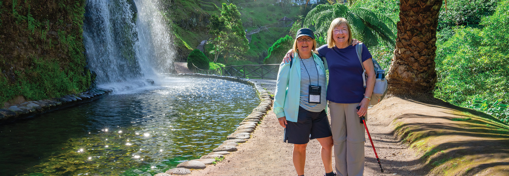 Two smiling women pause on a hiking trail next to a beautiful waterfall and lush green foliage in the Azores.