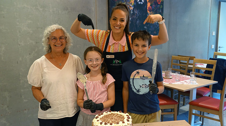 A group of four people, including two children, smile proudly behind a freshly made Black Forest cake during a workshop in Germany.