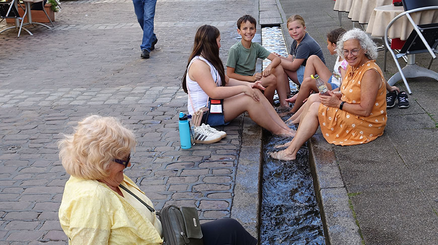 A group of adults and children cool their feet in a Freiburger Bächle, a small water channel running through a cobblestone street in Germany.