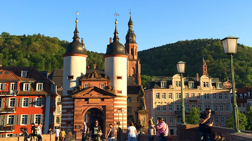 The twin-towered Old Bridge gate in Heidelberg, Germany, with people walking by, colorful buildings, and a forested hill in the background.