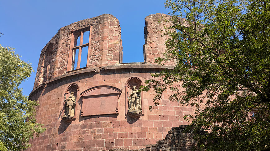 The crumbling red stone tower of Heidelberg Castle in Germany stands against a bright blue sky, framed by green leafy trees.