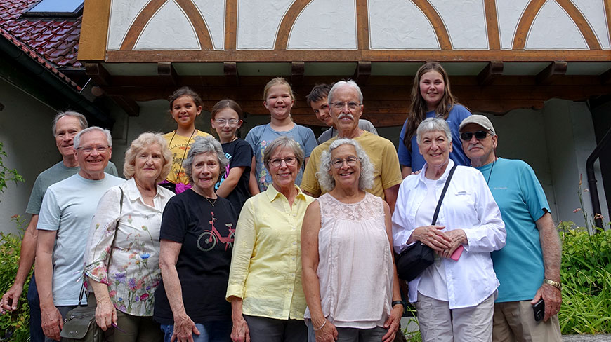 An intergenerational tour group smiles for a portrait in front of a timber-framed building in Triberg, Germany.