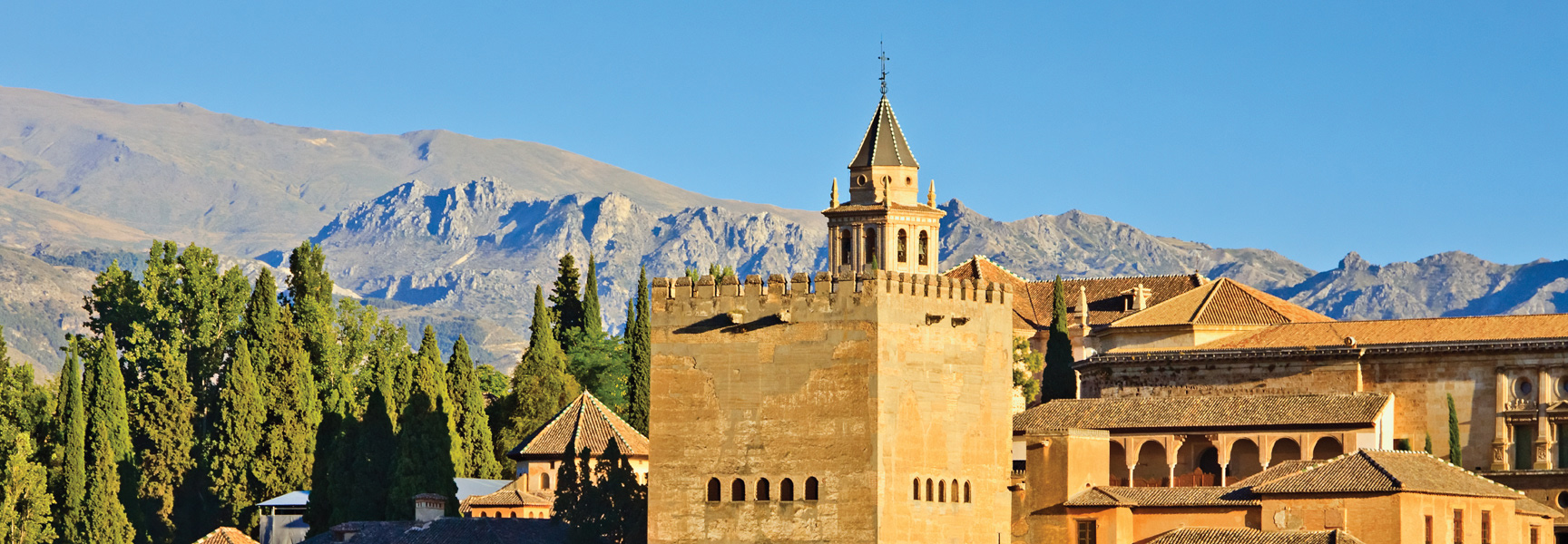 A sunlit, historic stone fortress stands before a rugged mountain range under a clear blue sky in Andalusia, Spain.